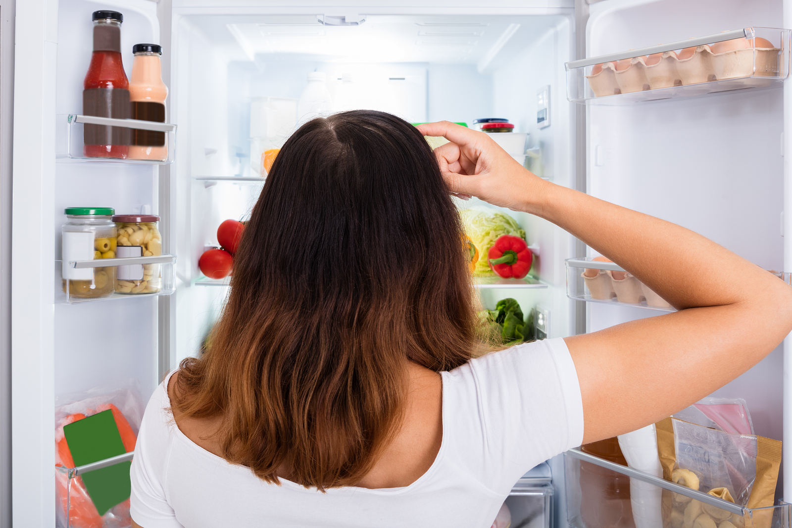 Rear View Of Confused Woman Searching For Food In The Fridge