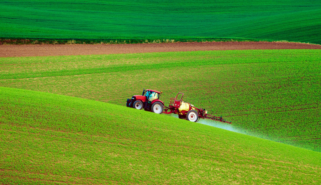 Farm machinery spraying insecticide drives up a lush, green, rolling hill in spring