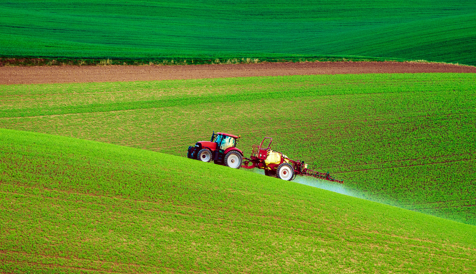Farm machinery spraying insecticide drives up a lush, green, rolling hill in spring