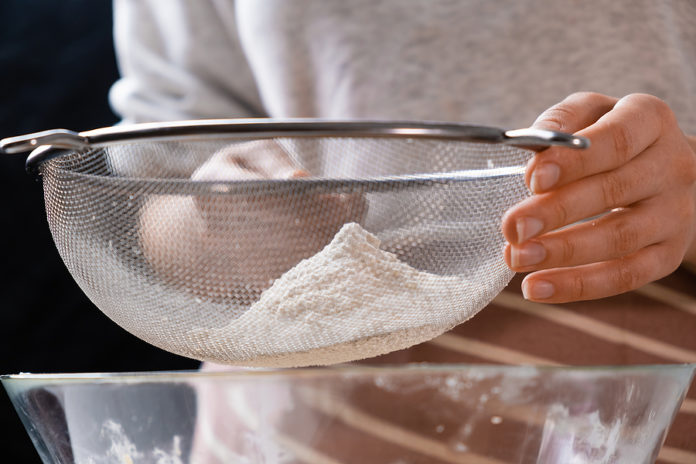 Close Up Woman Sifts Wheat Flour Through A Sieve Into Mixing Bow