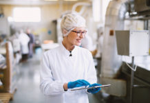 From Compliance to Competitive Advantage: Q&A With SafetyChain on the Real ROI of Digital Food Safety Smiley female worker in sterile clothes using tablet and checking how production line is working.