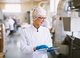 From Compliance to Competitive Advantage: Q&A With SafetyChain on the Real ROI of Digital Food Safety Smiley female worker in sterile clothes using tablet and checking how production line is working.