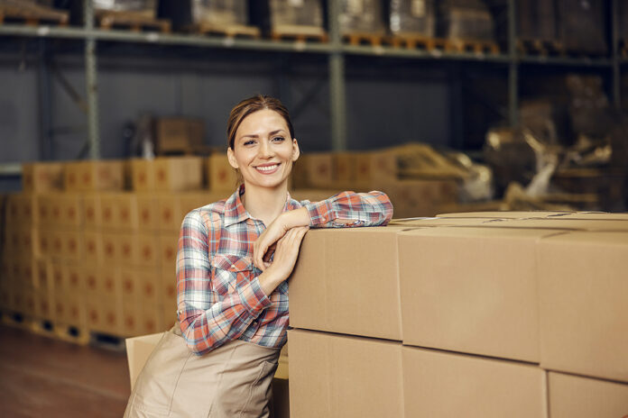 A food factory worker leaning on boxes in warehouse and smiling at the camera.