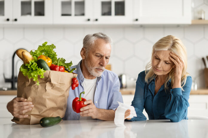 Frustrated Senior Spouses In Kitchen Checking Bills After Grocery Shopping